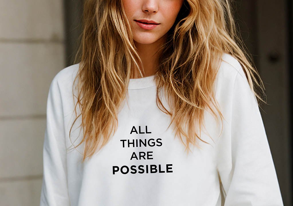 Close-up of a young blonde woman wearing an oversized organic cotton sweatshirt that features the positive quote, "All Things Are Possible."
