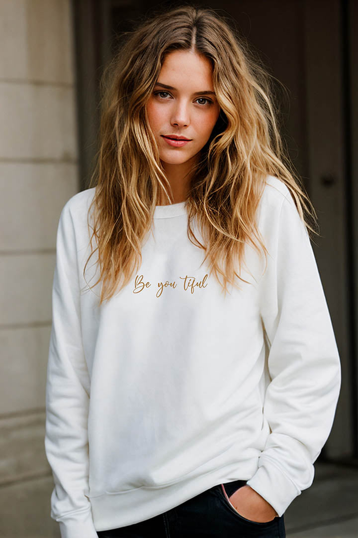 Upper body shot of a young blonde woman wearing an oversized organic cotton sweatshirt that features the positive saying, "Be you tiful."