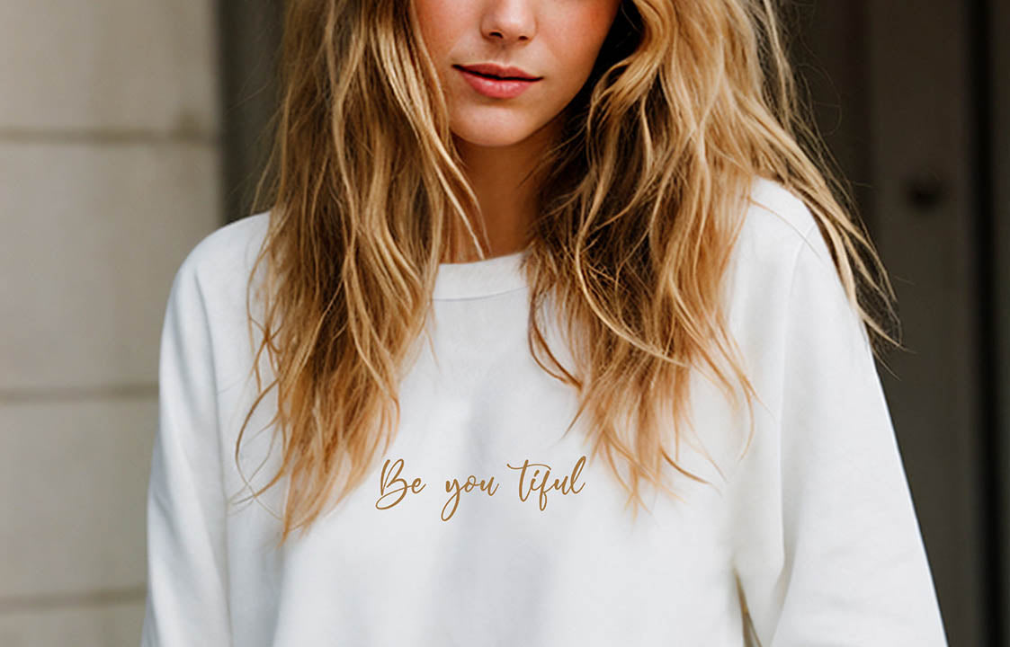 Close-up of a young blonde woman wearing a white organic cotton sweatshirt that features the positive saying, "Be you tiful."