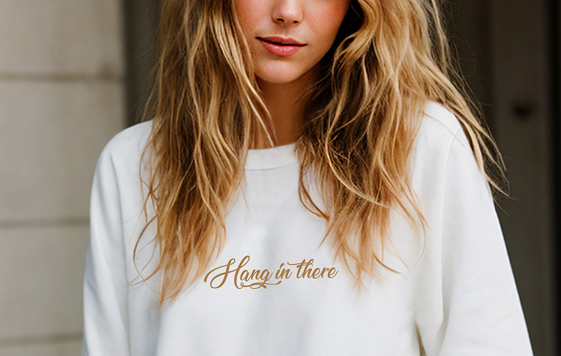 Close-up of a young blonde woman wearing an oversized organic cotton sweatshirt that features the positive quote, "Hang in there," in front of a neoclassical wall