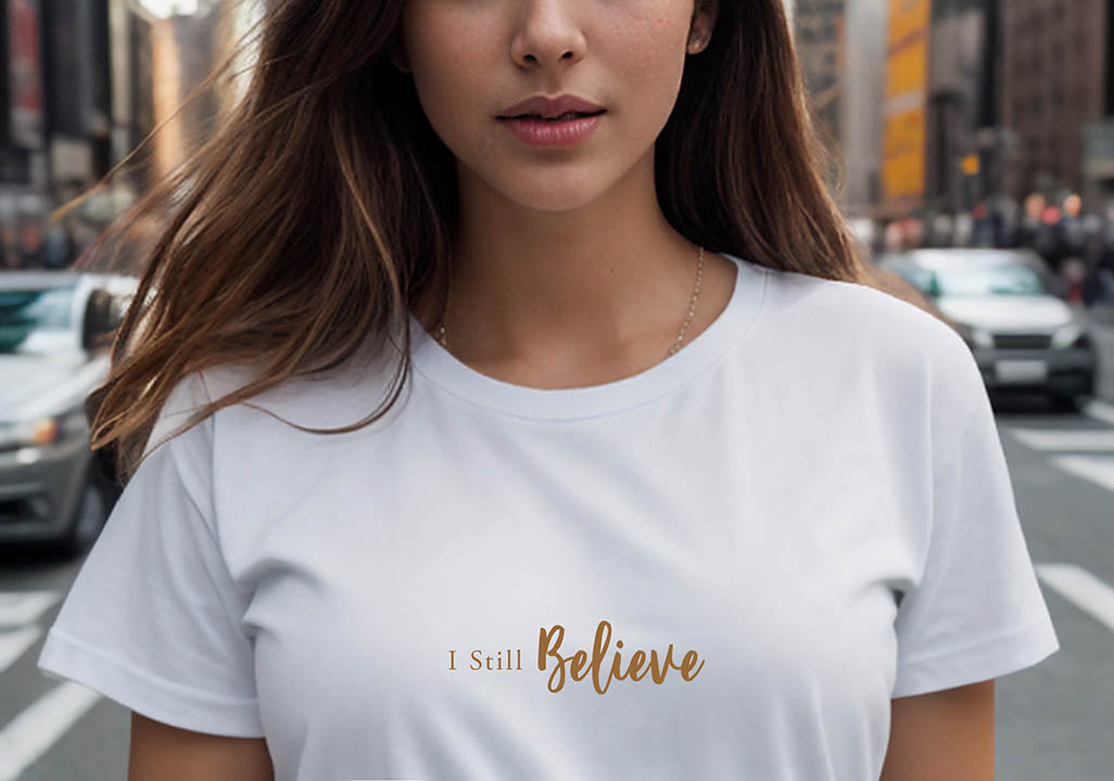 Close-up of a young brunette woman wearing a white 100% organic cotton t-shirt that features the positive message, "I Still Believe."