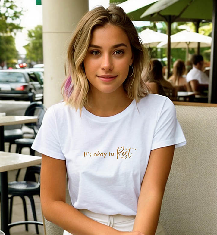 Inside a cafe a young blonde woman wearing a white 100% organic cotton t-shirt that features the positive quote, "It's okay to REST."