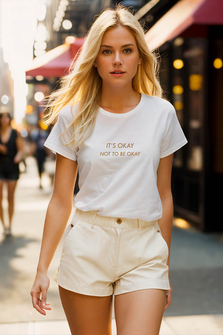 On a busy street a young blonde woman wearing a white 100% organic cotton t-shirt that features the positive quote, "It's okay to not be okay."