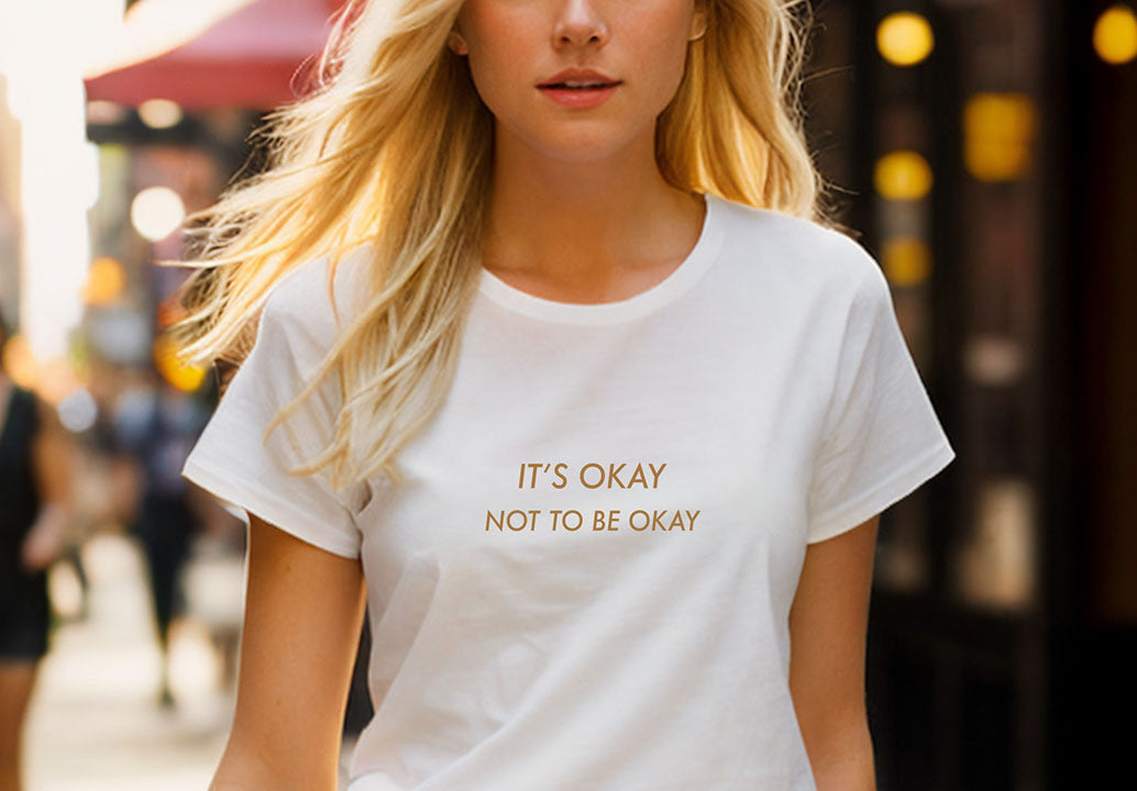 Close-up of a young blonde woman wearing a white 100% organic cotton t-shirt that features the positive quote, "It's okay to not be okay."