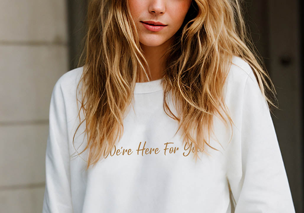 Close-up of a young blonde woman wearing an oversized organic cotton sweatshirt that features the positive quote, "We're Here For You," in front of a neoclassical wall