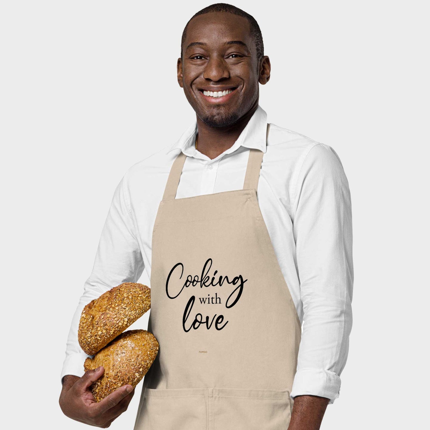 Man wearing a beige "Cooking with Love" inspirational quote 100% organic cotton apron, his right hand holding breads
